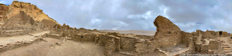 A narrow path passes between low stone walls toward a tall, slender standing masonry remnant.