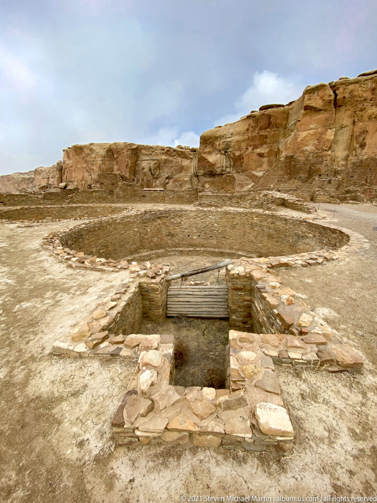 A rectangular stone-lined opening with wooden beams sits before a broad circular enclosure and canyon wall.