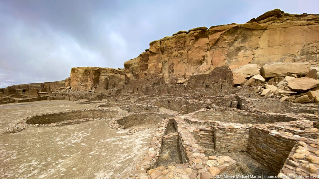 Circular and rectangular stone remains lie below a sandstone cliff, beside a broad perimeter path.