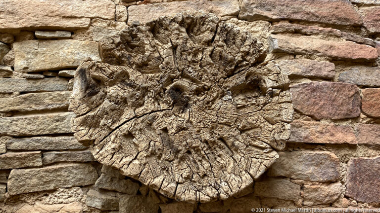 A cracked circular wood end is embedded in a sandstone wall among fitted stone blocks.