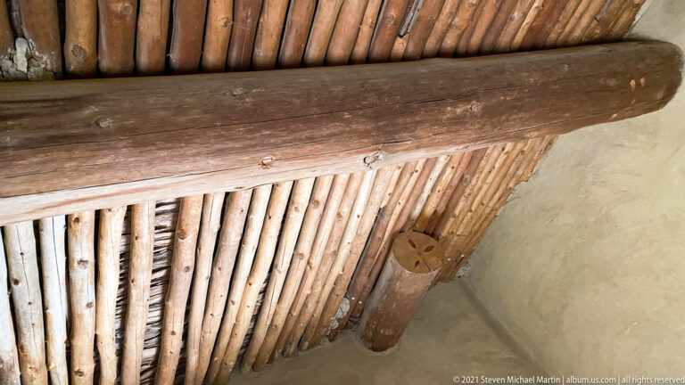 Parallel wooden poles form a ceiling beneath a thick crossbeam, viewed from below.
