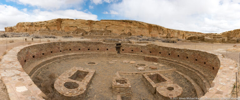 An elevated view looks into a circular kiva with low interior structures beneath a sandstone canyon wall.