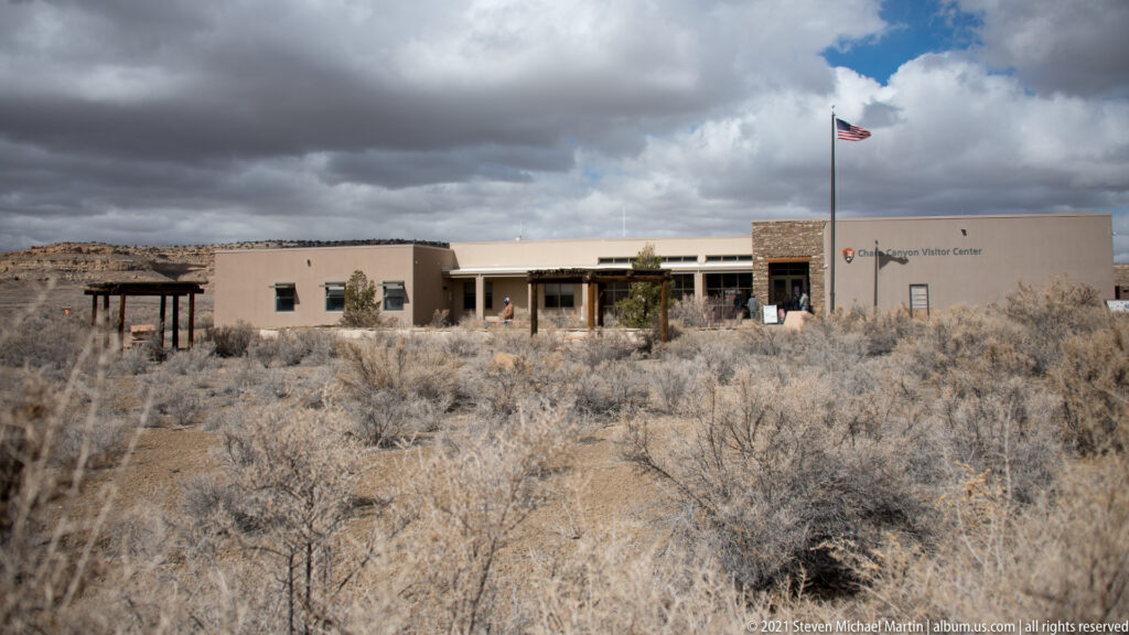 A low visitor center and raised American flag stand beyond sagebrush, with a mesa under cloudy skies.