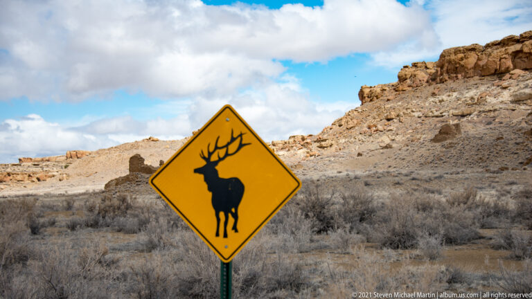 A yellow deer-crossing sign stands beside a gravel road with sandstone cliffs and scrub behind it.