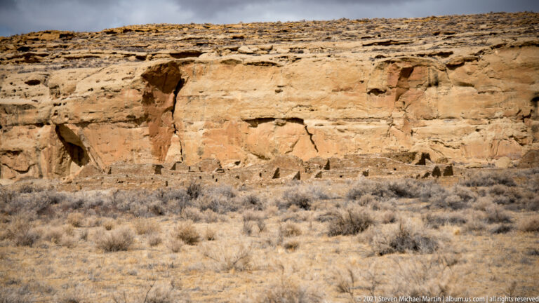 A low layered sandstone wall with a dark recess overlooks a slope of sparse desert shrubs.