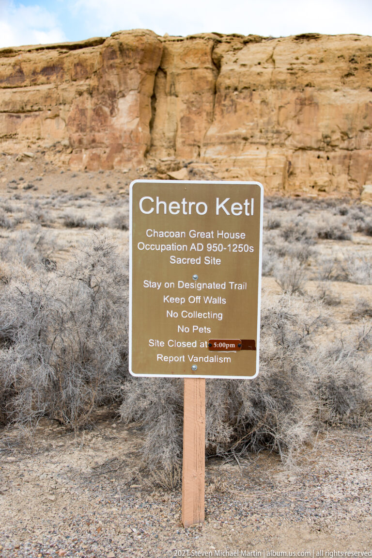 A brown Chetro Ketl sign stands in desert scrub before a sandstone cliff, identifying the Great House and trail rules.