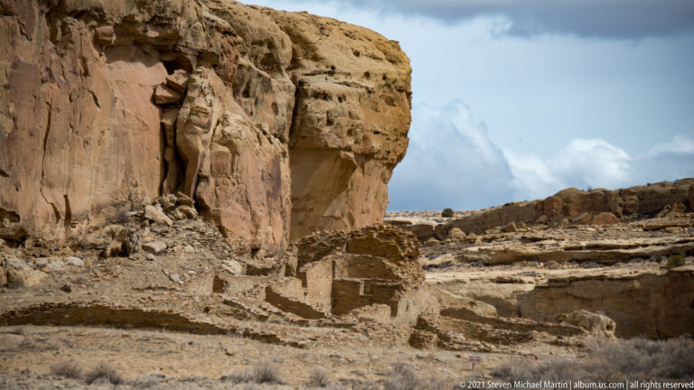 Low stone walls stand beneath a sheer sandstone cliff, with rubble and open ground in the foreground.