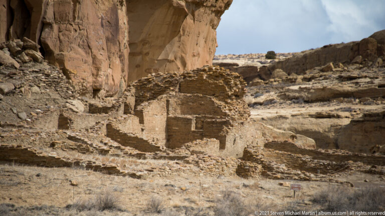 A low masonry room block lies beneath a projecting sandstone cliff, with a small trail marker nearby.