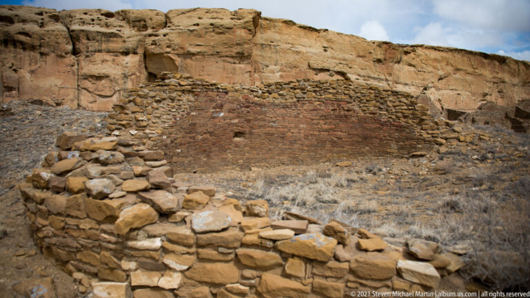 A low curved stone wall with broad capstones stands before a layered sandstone cliff.