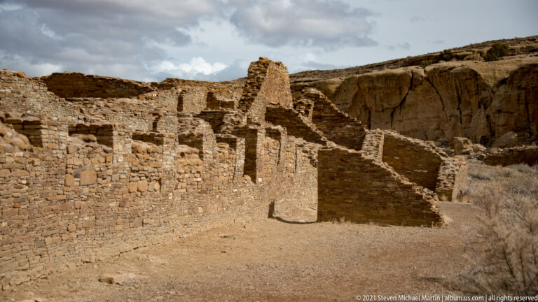 Multiple low stone walls and taller room blocks extend across the base of a sandstone cliff.
