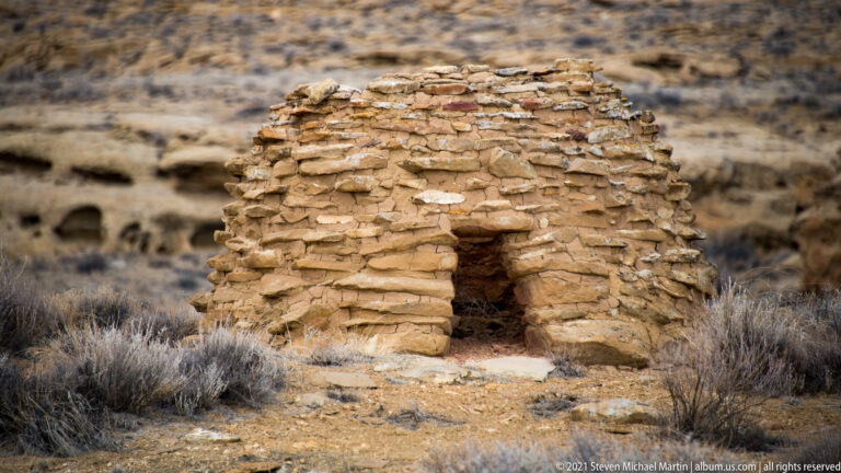 A small, partly collapsed stone wall stands among desert shrubs and scattered masonry fragments.