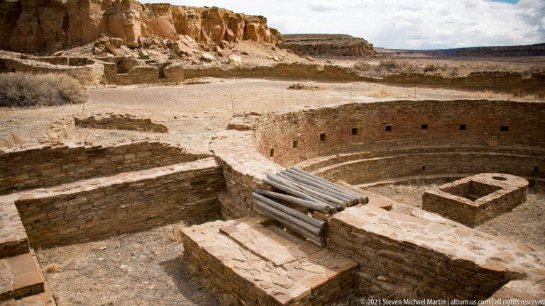 A circular sunken stone structure with curved interior walls and timber beams lies among nearby ruins.