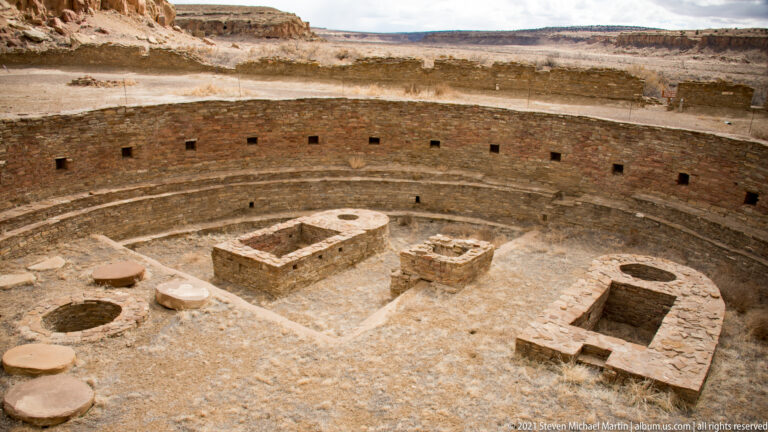 A large circular stone enclosure with interior rooms sits below a curved masonry wall in a sandstone canyon.