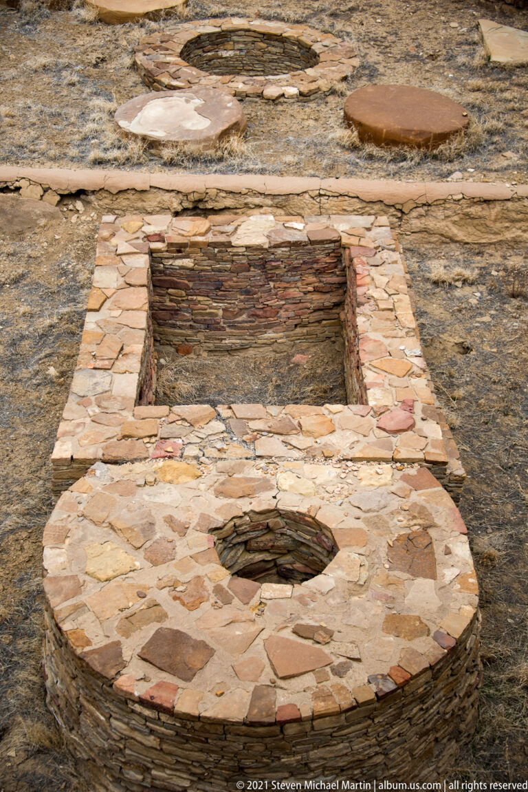 A rectangular stone-lined depression lies between two circular openings in a paved sandstone archaeological area.