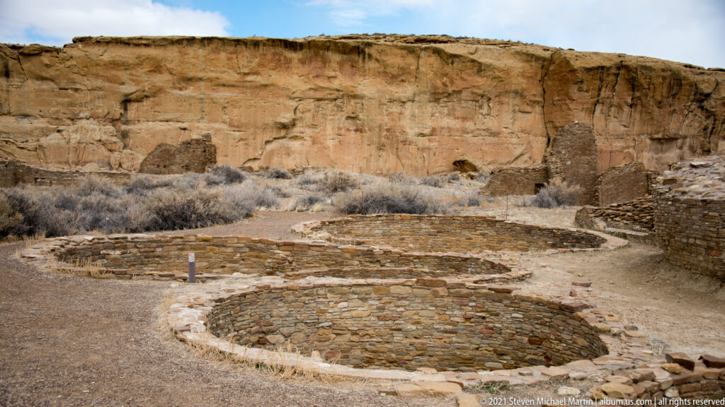 Circular stone enclosures and low masonry walls lie below a tall sandstone cliff.