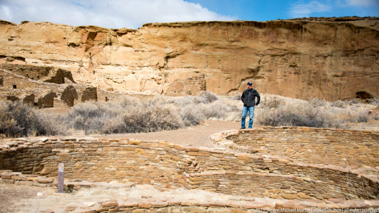 A visitor stands on a dirt path among low stone walls and circular foundations beneath a sandstone cliff.