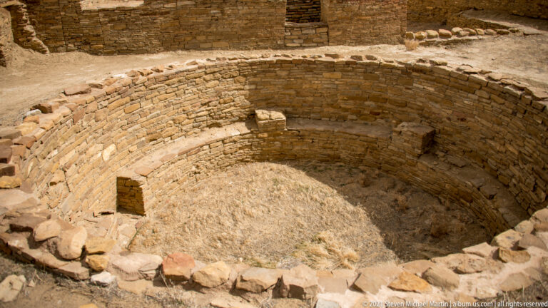 A deep circular stone enclosure has curved inner walls, a narrow ledge, and dry grass across its floor.