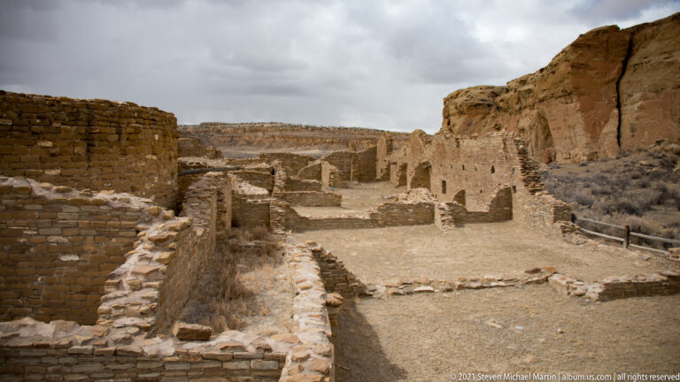 A path passes between stone room walls toward taller masonry ruins and sandstone cliffs.
