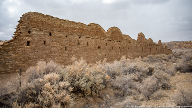 A long curved stone façade with small square openings extends beside dry brush beneath a cloudy sky.