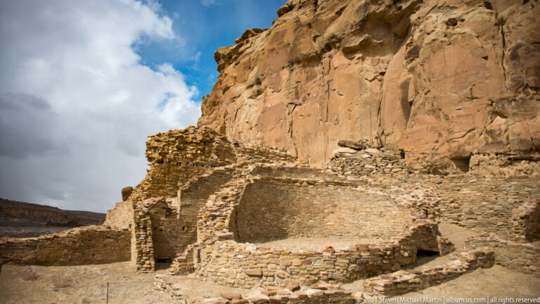 Masonry rooms and standing walls sit directly below a towering orange sandstone cliff.