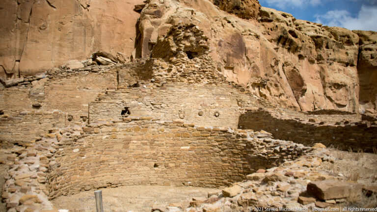 A broad stone façade with small openings and broken upper courses stands against a sandstone cliff.