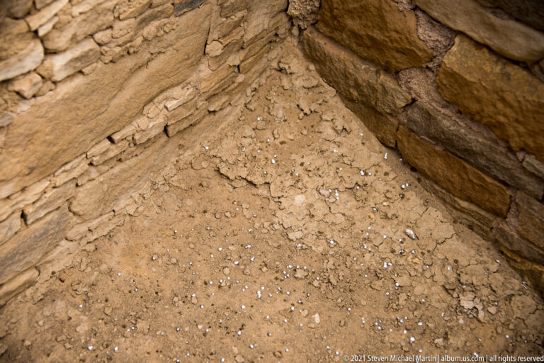 Two sandstone walls meet above a sloping dirt floor scattered with small stones.