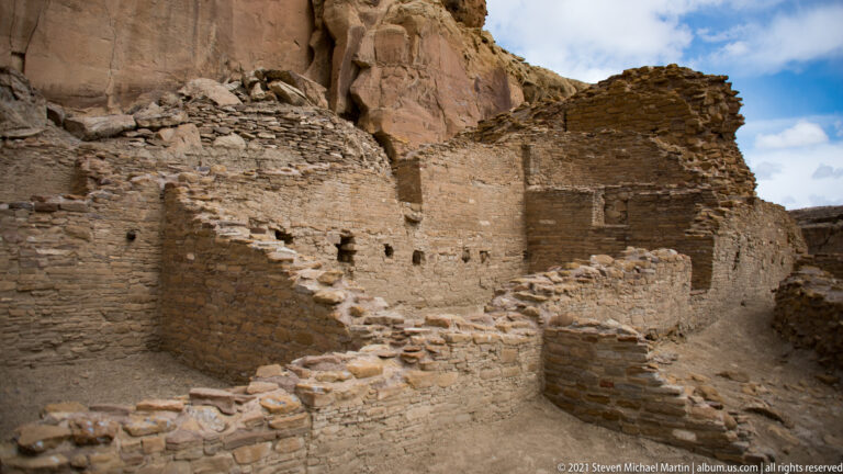 Uneven stone steps rise between masonry walls toward higher rooms beneath a sandstone cliff.