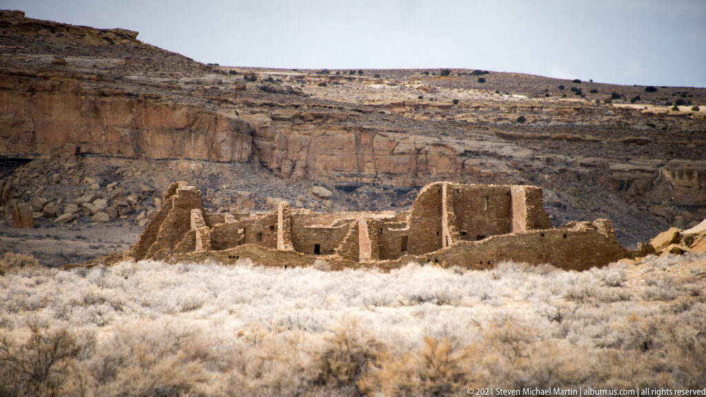 Low stone ruins stand across an open canyon floor below a long layered rim.