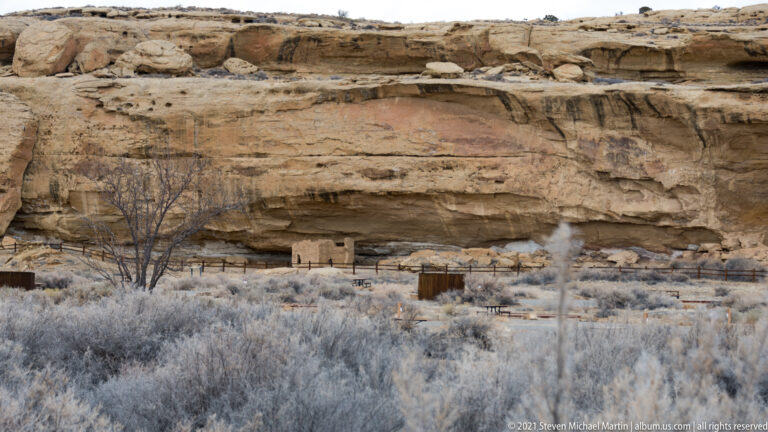 Small masonry structures sit beneath a wide sandstone overhang, beyond winter shrubs and a low fence.