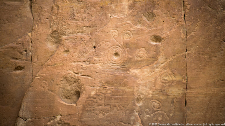 A weathered rock face bears a central ringed figure, small spirals, circles, and other pecked marks.