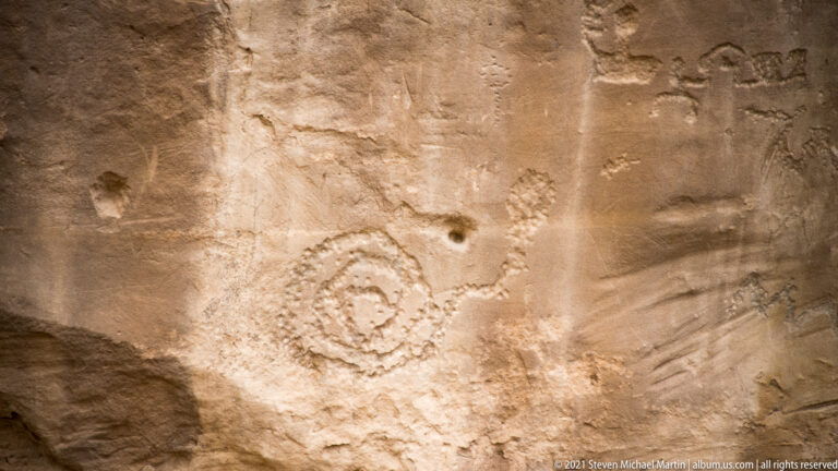 A large ringed spiral-like carving appears on pale sandstone amid faint surrounding marks.