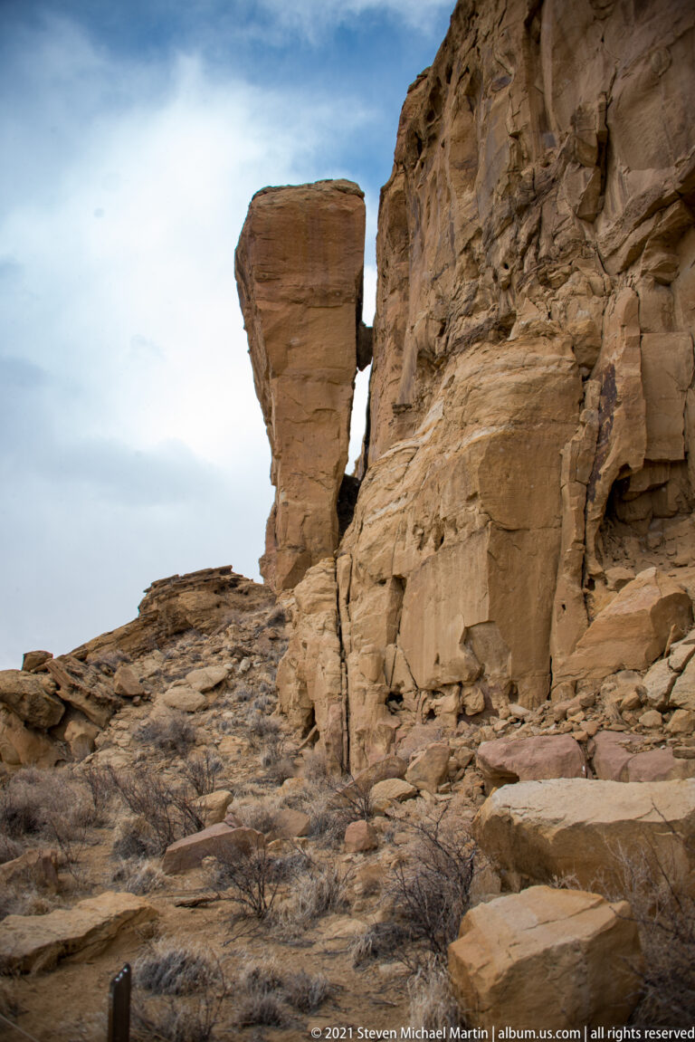 A tall block-shaped sandstone outcrop rises above broken rock and low ruins beside a cliff.