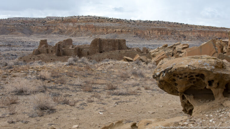 A distant masonry ruin stands across a dry canyon floor, framed by a large pitted boulder at right.