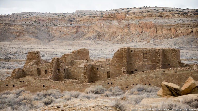 Rectangular masonry rooms with broken upper walls stand below a long canyon rim.