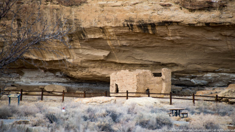 A rectangular stone ruin stands behind a low barrier beneath a broad sandstone overhang.
