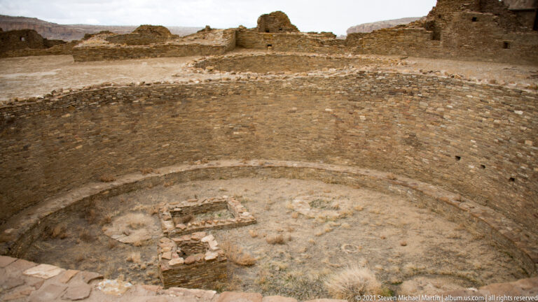A circular stone room contains a curved inner ledge and low rectangular foundations on a sandy floor.