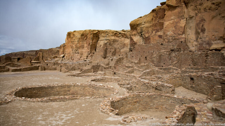Two circular stone-lined structures sit before a group of room walls and a tall cliff.
