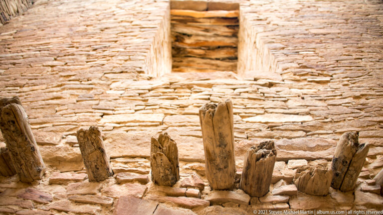 Upright wooden posts stand in a curved stone chamber beneath a small raised wall opening.