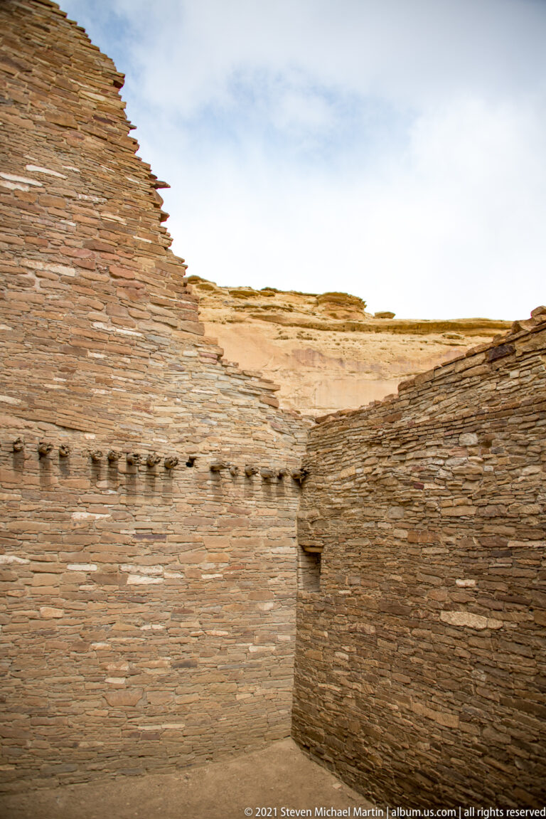 An open-topped stone enclosure has a sandy floor, a row of square wall holes, and a narrow doorway.