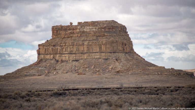 A flat-topped sandstone butte stands alone on a broad, sparsely vegetated plain beneath clouds.