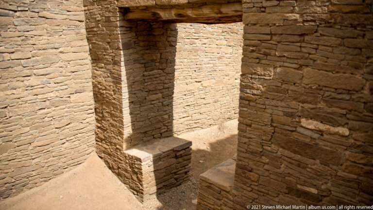 A sunlit stone chamber has thick walls, a deep doorway, and low ledges along the floor.