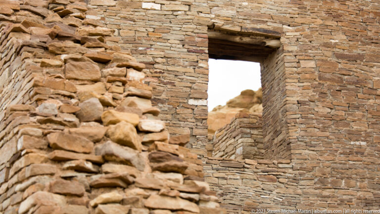 A narrow doorway cuts through a high stone wall beside fallen rubble, with a pale rock formation beyond.