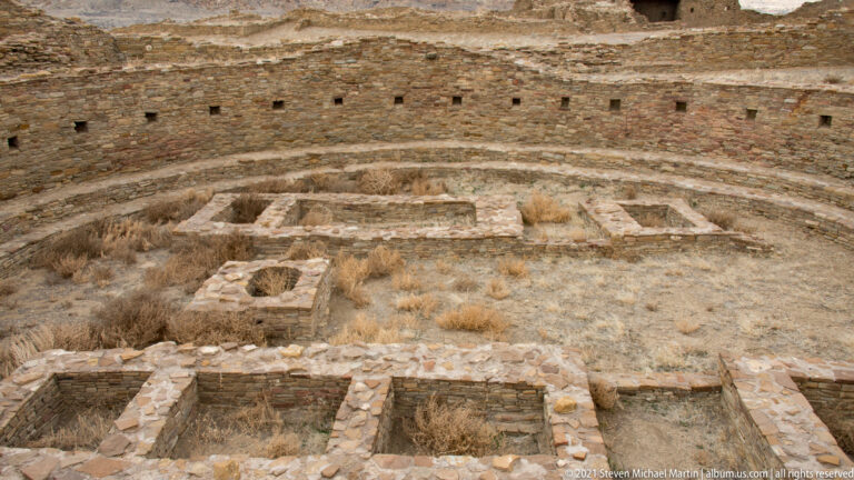 A circular stone chamber contains low rectangular compartments and a raised central feature beneath a curved wall.