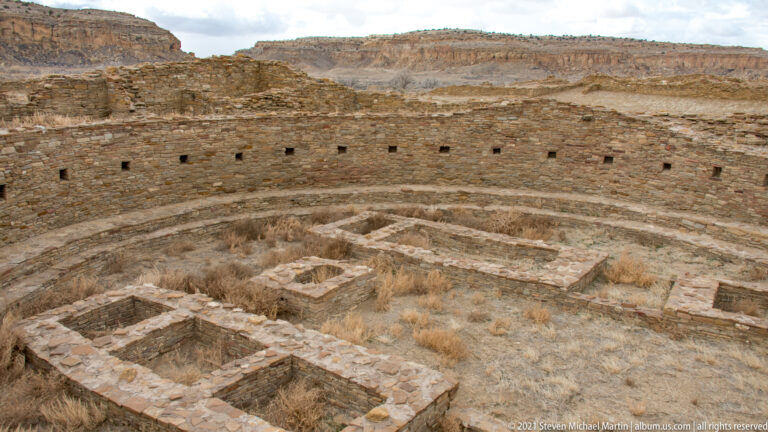 A wide circular stone room has small wall openings, a curved ledge, and rectangular compartments across its floor.