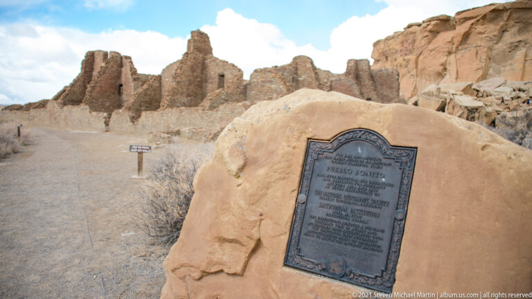 A metal plaque on a sandstone boulder stands before low stone ruins and a high rock wall.