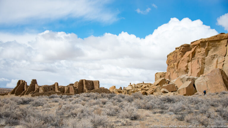 Low stone ruins and a visitor sit below a tall sandstone cliff beneath a clouded sky.