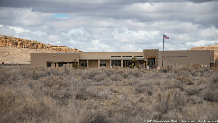 A low visitor center with a flagpole stands beyond sagebrush, backed by sandstone cliffs and cloudy sky.