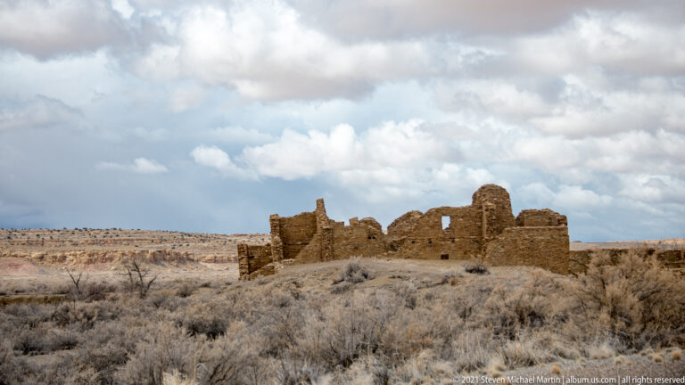 Broken stone walls stand alone on a scrub-covered rise beneath a cloudy sky, with distant mesas behind.