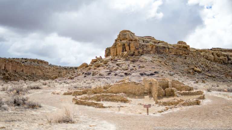 Low stone foundations sit below a rugged sandstone rise, with a small sign and scrub nearby.