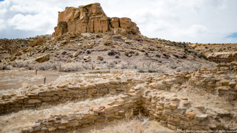 Stone foundations and broken walls lie below a large sandstone butte, with shrubs and rubble between them.